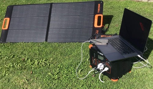Unbranded 100W solar panel on grass connected to a portable power station charging a laptop in full sun