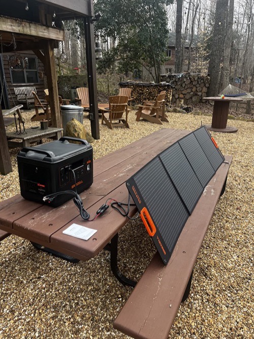 Unbranded 100W portable solar panel propped on a picnic table connected to a power station in a wooded backyard