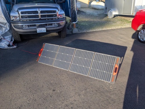 Unbranded 100W portable solar panel laid flat on an asphalt driveway next to a pickup truck