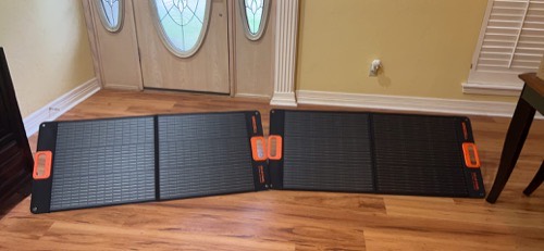 Two unbranded 100W foldable solar panels unfolded on a hardwood floor indoors near a front door