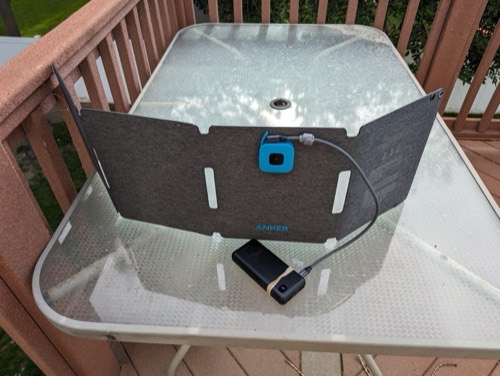 Anker SOLIX PS30 solar panel propped on a deck table with a USB-C cable running from the blue junction box to a power bank in the sun