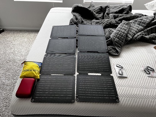 Two Anker SOLIX PS30 solar panels partially unfolded on a bed alongside a first aid kit, USB cables, and carabiners during unboxing