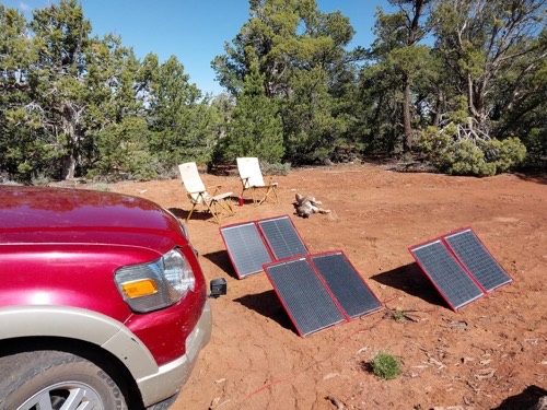 Two DOKIO 100W folding solar panel arrays deployed on red desert dirt beside a maroon SUV at a remote campsite