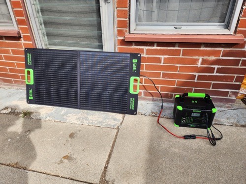 EBL 100W portable solar panel unfolded on a patio against a brick wall, charging an EBL power station