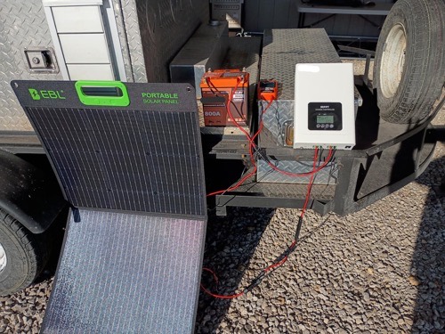 EBL 200W solar panel leaning against a truck wired through a charge controller to a 12V battery on gravel