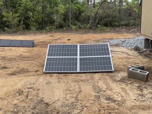Two ECO-WORTHY 400W solar panels ground-mounted on dirt beside an off-grid building, with a third panel folded nearby