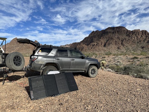 EcoFlow 220W bifacial solar panel deployed on the ground beside an SUV at a desert campsite under a blue sky