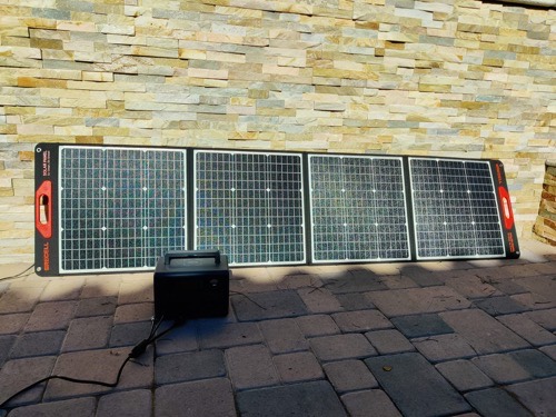 GRECELL SP-100 100W folding solar panel with red handles unfolded against a stone wall, feeding a black power station