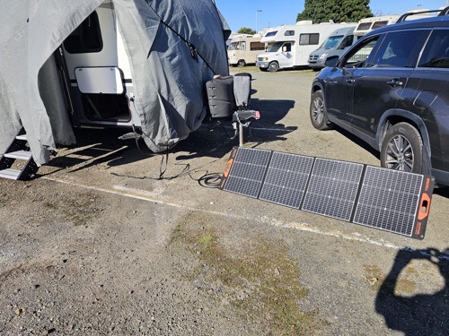 GRECELL SP-100 folding solar panel deployed on the ground at an RV campsite next to a travel trailer and SUV