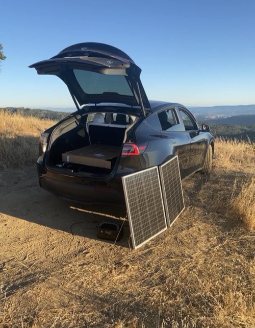Two HQST 200W bifacial solar panels propped against the open hatch of a black SUV charging a power station on a hilltop