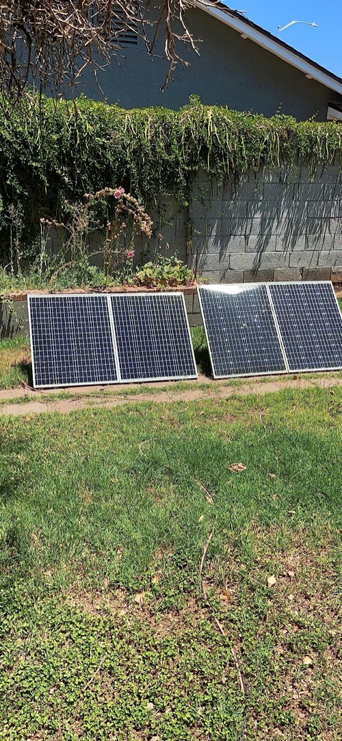 Two HQST bifacial 100W solar panels leaning on simple ground stands on grass against a garden wall in full sun