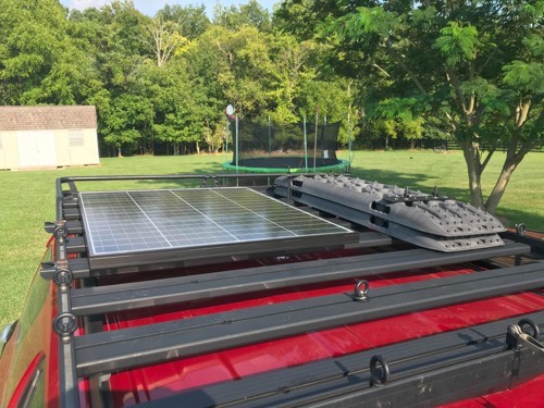 HQST bifacial 100W solar panel strapped to a vehicle roof rack next to recovery traction boards in a grassy yard