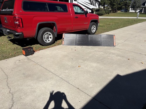 Jackery SolarSaga 200W solar panel laid out on a driveway beside a red pickup truck with an orange Jackery power station and an RV in the background