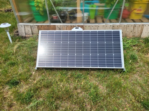 Renogy 100W 12V monocrystalline solar panel leaning against a greenhouse on grass