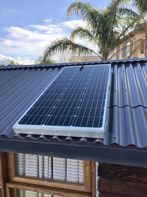 Renogy 100W 12V solar panel mounted on a corrugated metal roof with Z-brackets under palm trees