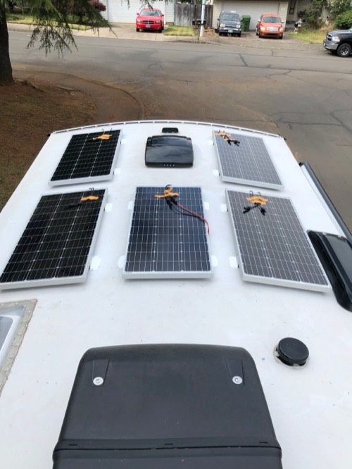 Six Renogy 100W 12V solar panels laid out across a van roof in a driveway during install