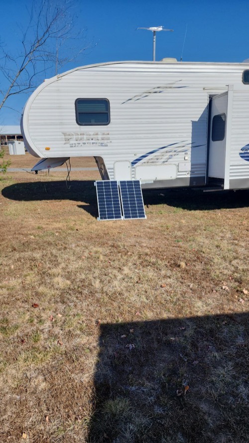 Two Renogy 100W solar panels leaning against a fifth-wheel trailer on dry grass in full sun
