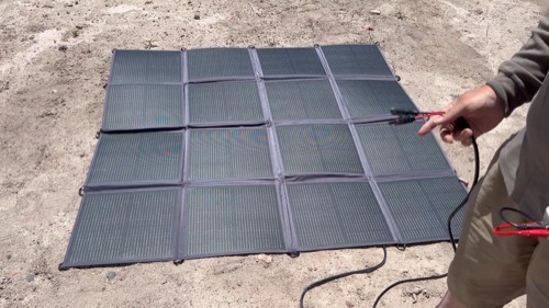 Owner holding the Renogy 400W solar blanket MC4 leads with multimeter probes to test output, panel laid flat on dirt in full sun