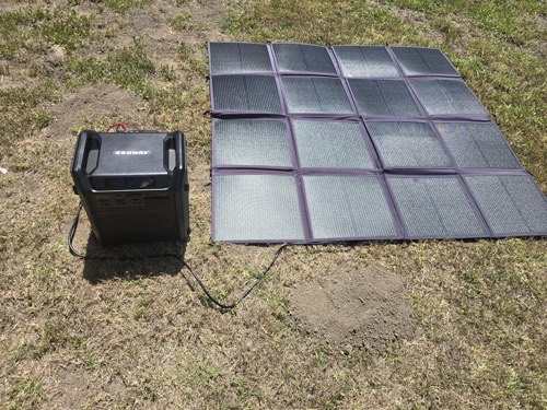 Renogy 400W solar panel blanket laid flat on grass connected by cable to a portable power station charging in the sun