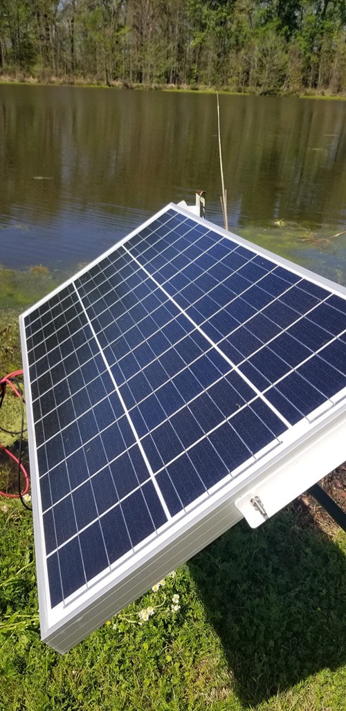 Topsolar 100W monocrystalline solar panel mounted outdoors on a tilt frame beside a pond, charging a 12V battery off-grid
