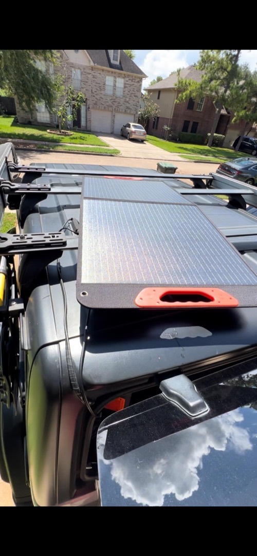 ZOUPW 100W solar panel laid flat across a vehicle roof rack in a suburban driveway to capture midday sun